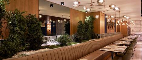 A long, modern dining area with a central bench, multiple tables, and ambient lighting.