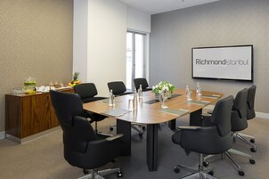 A conference room with a long table, black chairs, a wooden desk with a plant, and a screen displaying "Richmondistanbul".