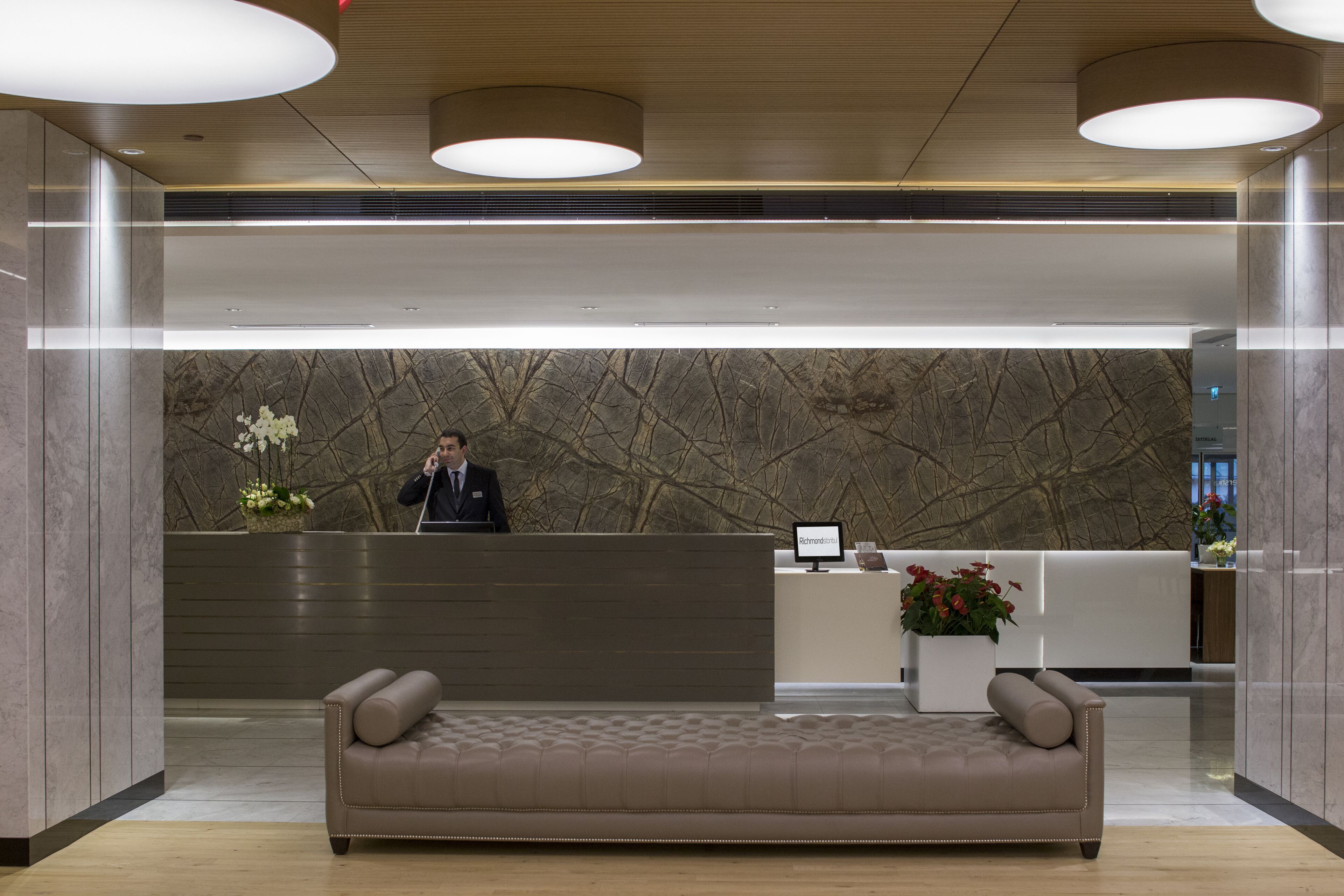 A reception area with a marble wall, a receptionist, a sofa, and a potted plant.