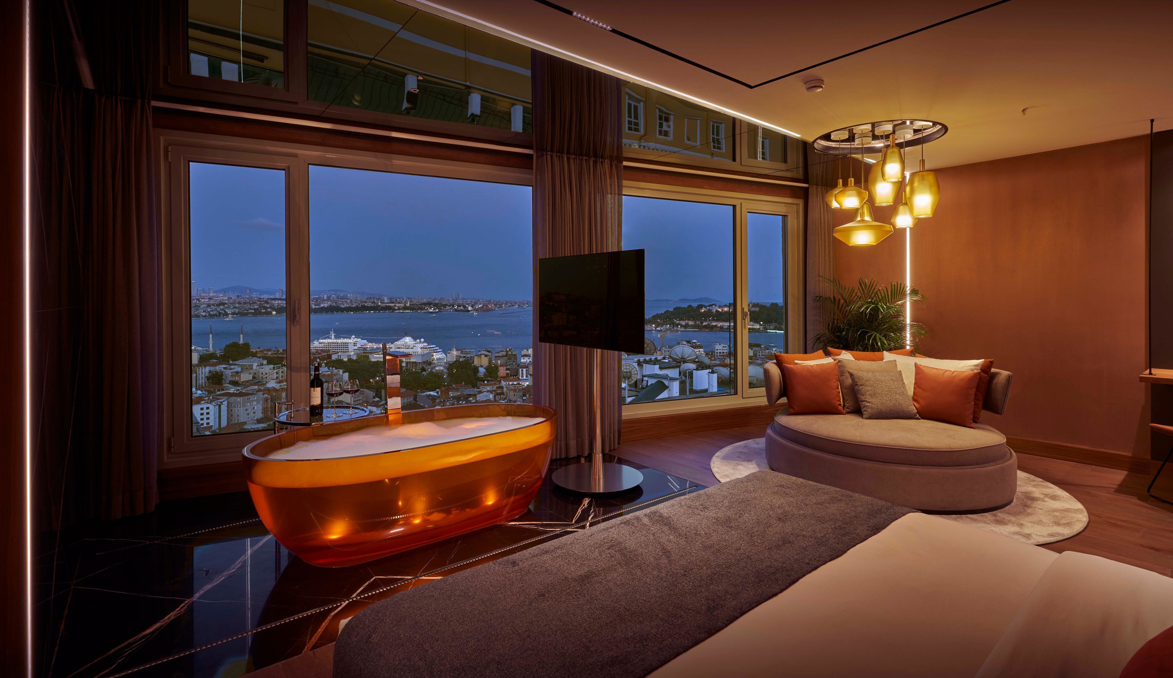 A modern hotel room with a large window offering a city view, a bathtub, a sofa, and a flat-screen TV.