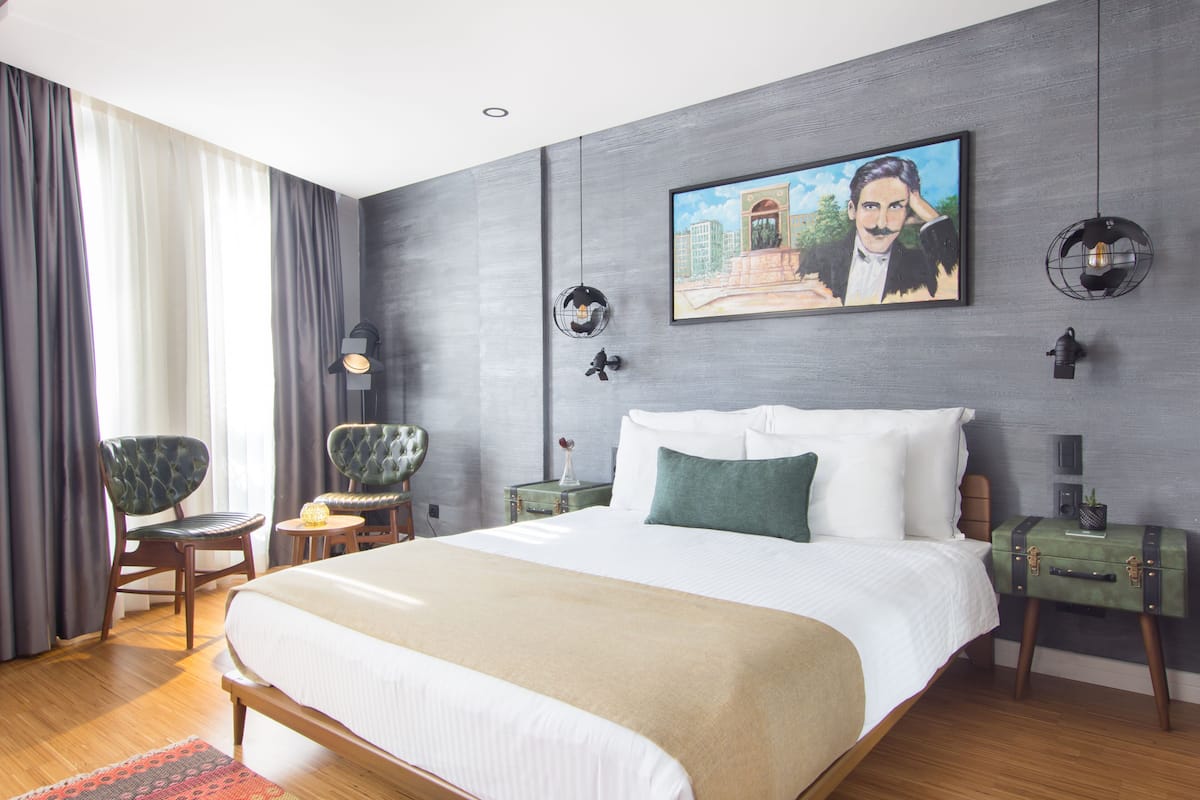 A modern bedroom with a large bed, a painting on the wall, and a view of the outdoors through the window.