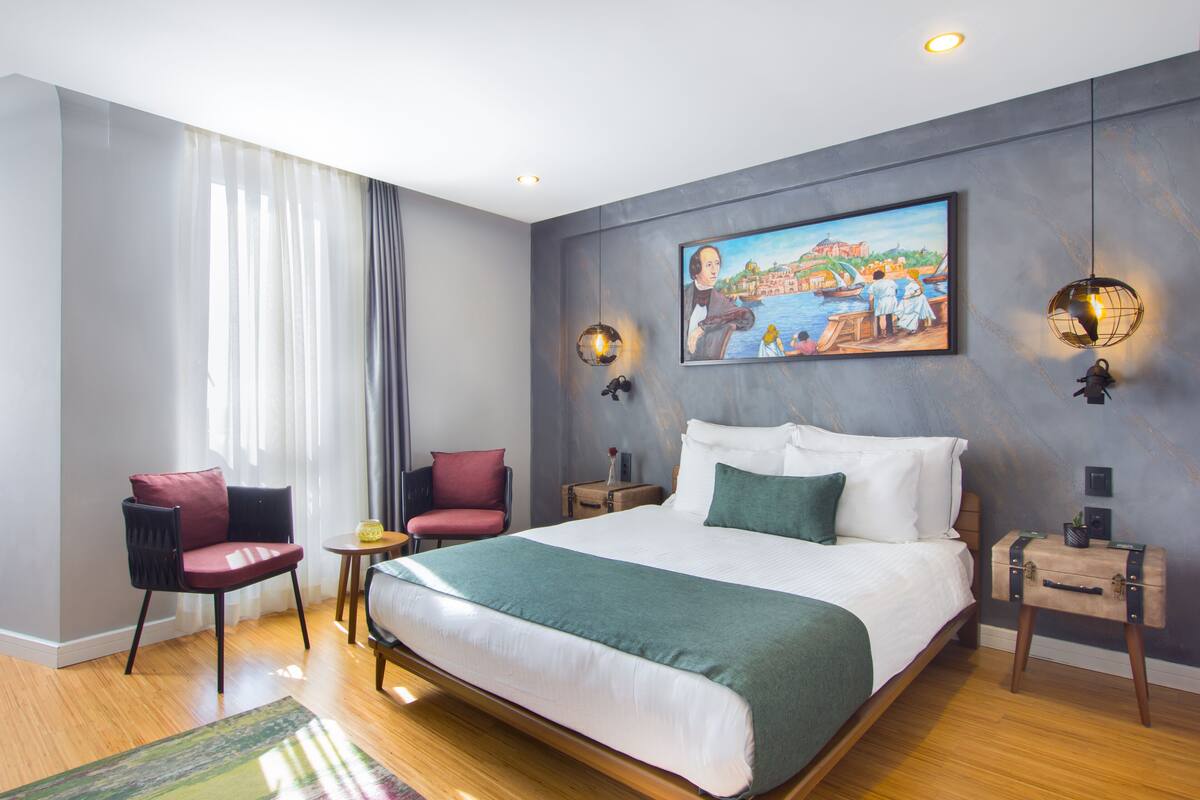 A modern hotel room with a large bed, two chairs, a small table, and a painting on the wall.