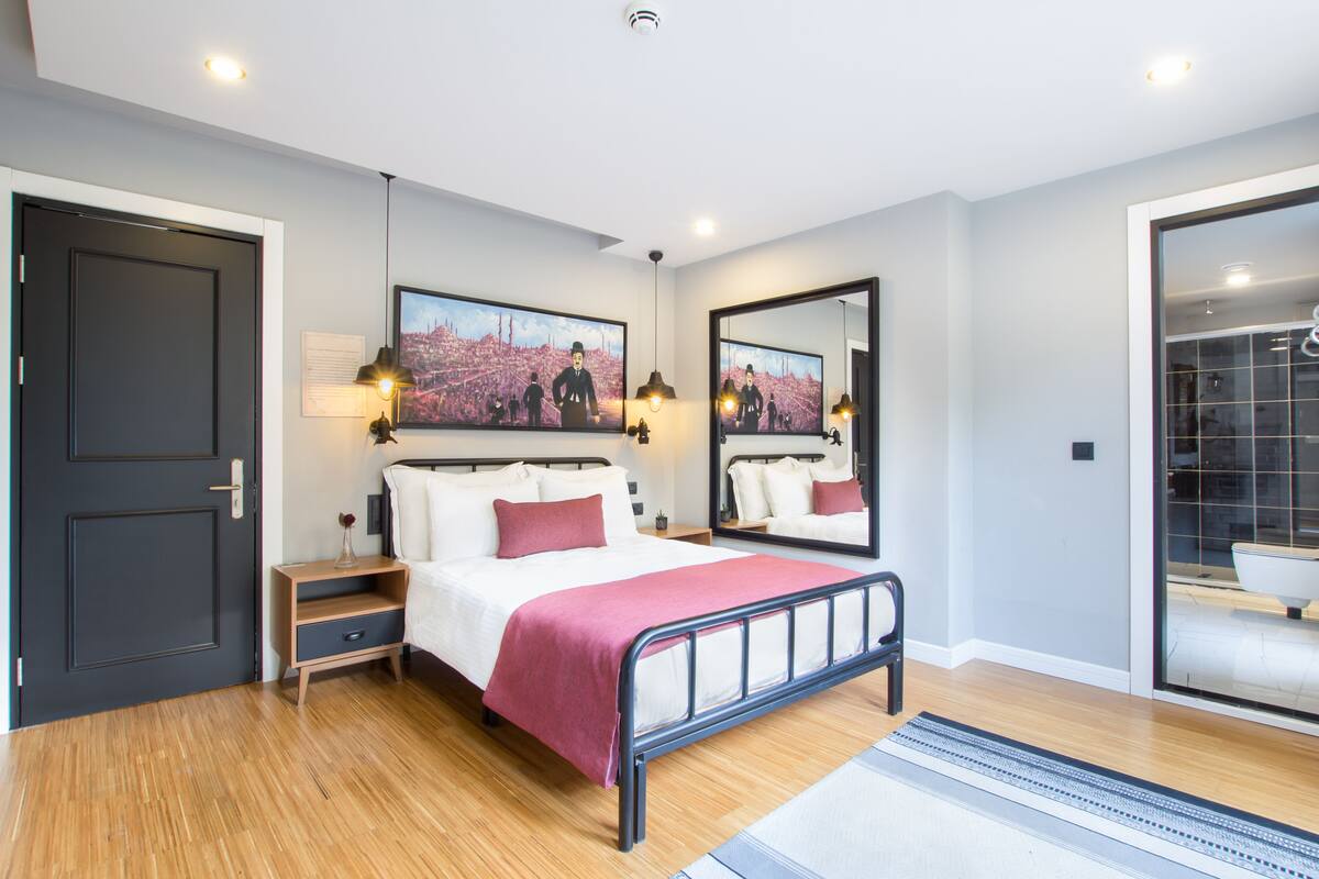 A modern bedroom with a bed, bedside tables, a mirror, and a cityscape painting on the wall.