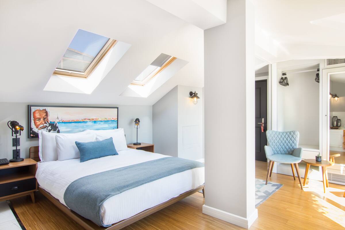 A bedroom with a large bed, skylights, a painting, a chair, and a small table.