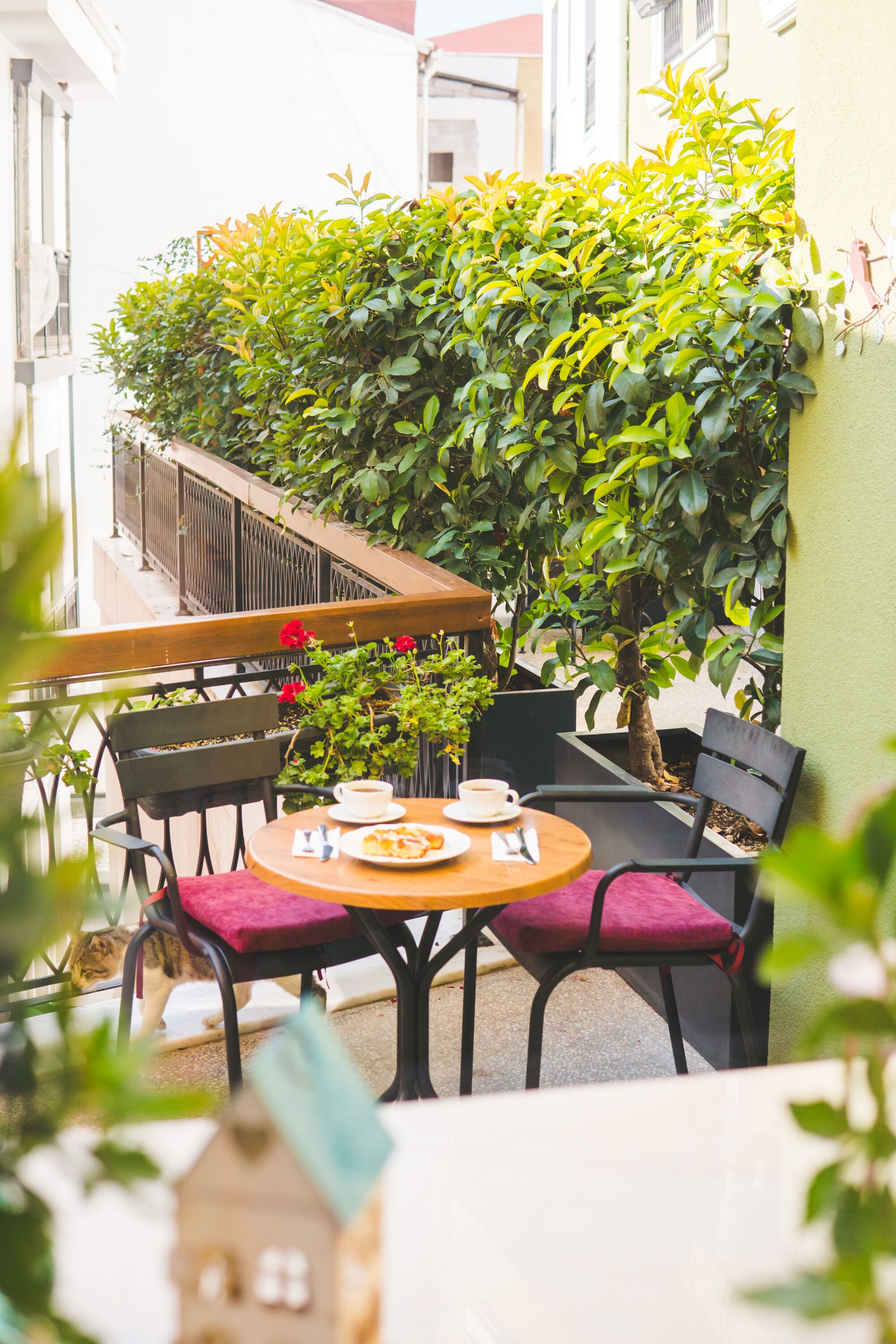 A small outdoor seating area with a table and chairs, surrounded by greenery and a railing.