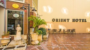 Un espace extérieur aménagé avec des chaises et une statue, une enseigne indiquant l’hôtel Orient, et une plante en pot.