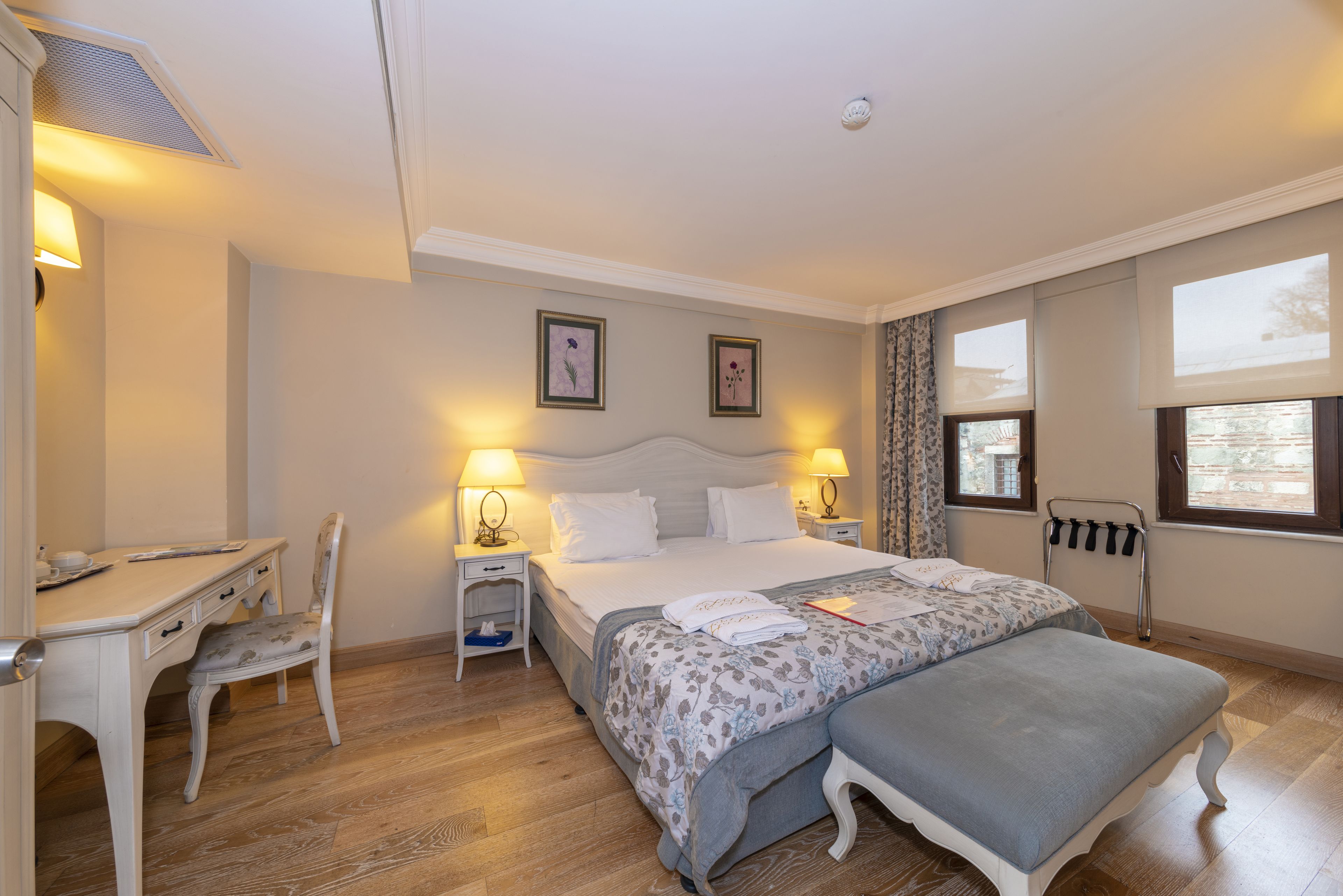 A hotel room with a bed, bedside tables, a desk, a chair, and a window.