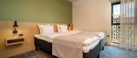 A hotel room with a bed, bedside table, lamp, and a view of the outside.