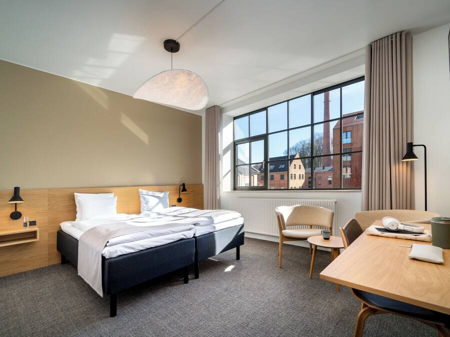 Una camera d'albergo con un letto, una scrivania, una sedia, una finestra vista edifici e una lampada a sospensione.