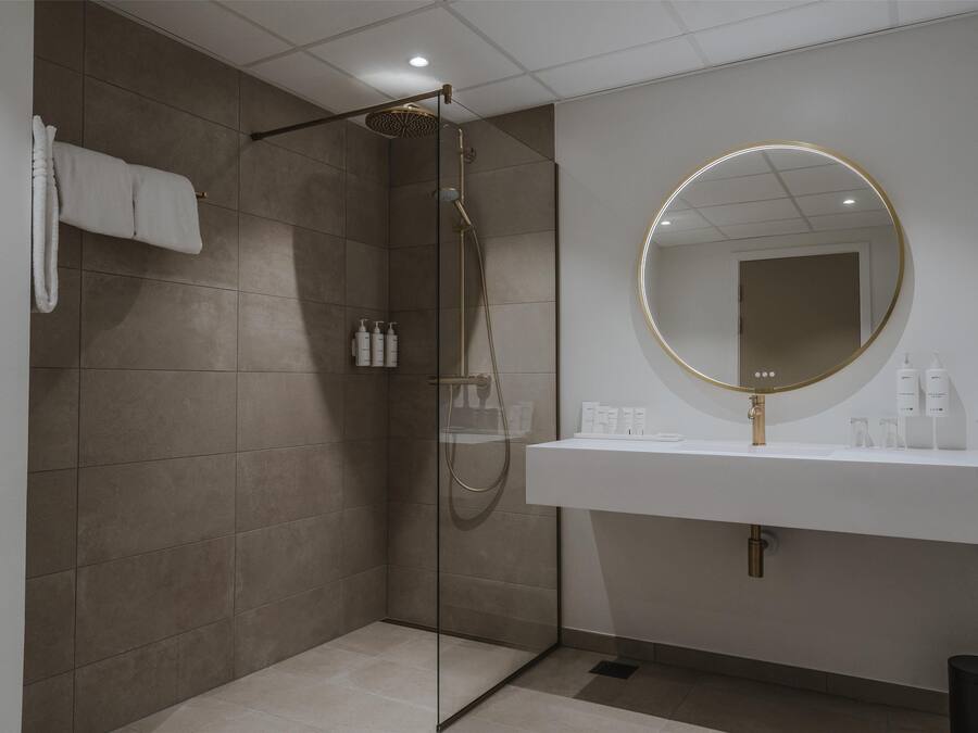 A modern bathroom with a glass-enclosed shower, a white sink with a gold mirror, and toiletries on the counter.