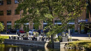 Un patio a la orilla del río con mesas al aire libre, un hotel Radisson al fondo y una masa de agua tranquila.