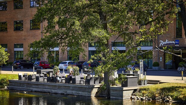 Un patio a la orilla del río con mesas al aire libre, un hotel Radisson al fondo y una masa de agua tranquila.