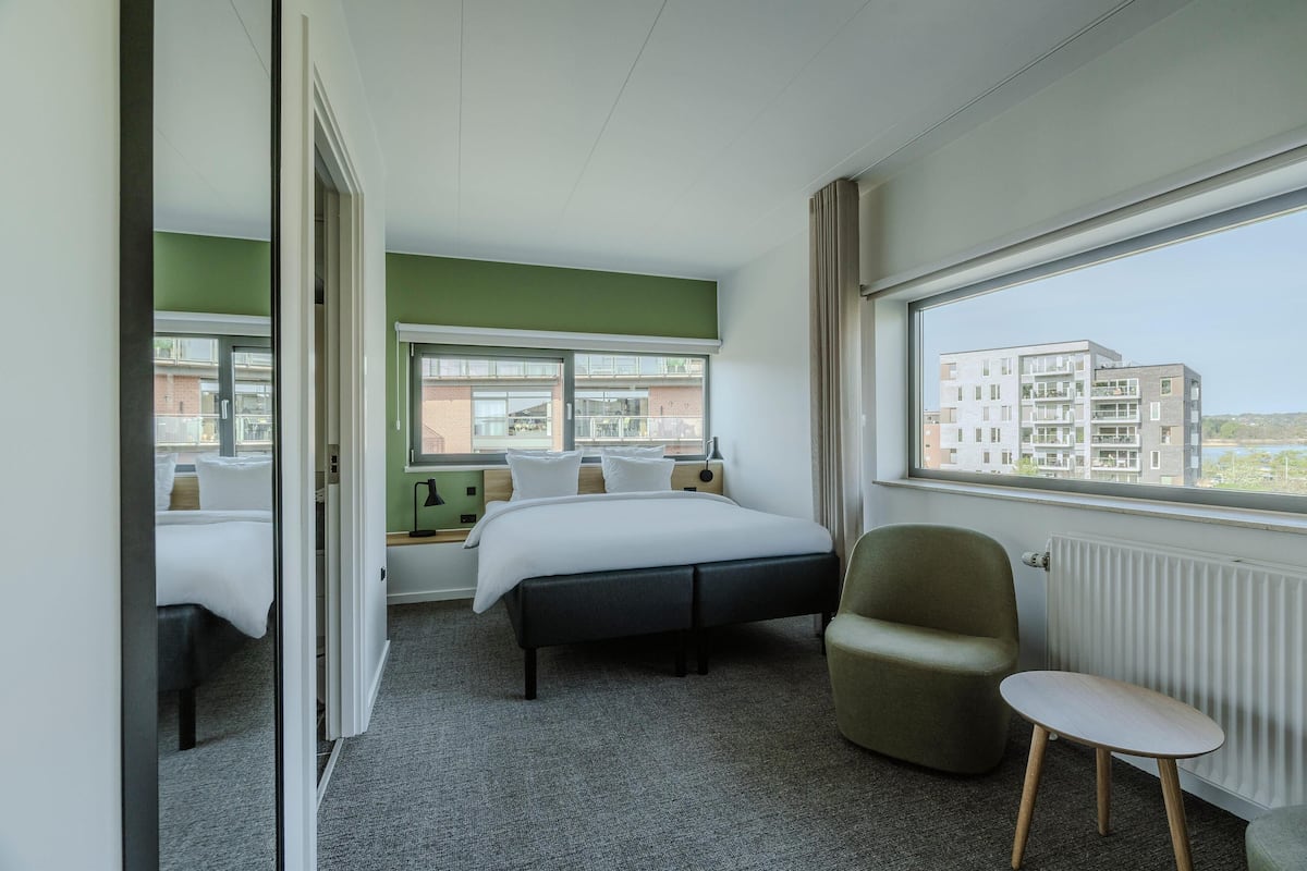 Et hotelværelse med en seng, en stol, et lille bord og et stort vindue med udsigt til bygninger.