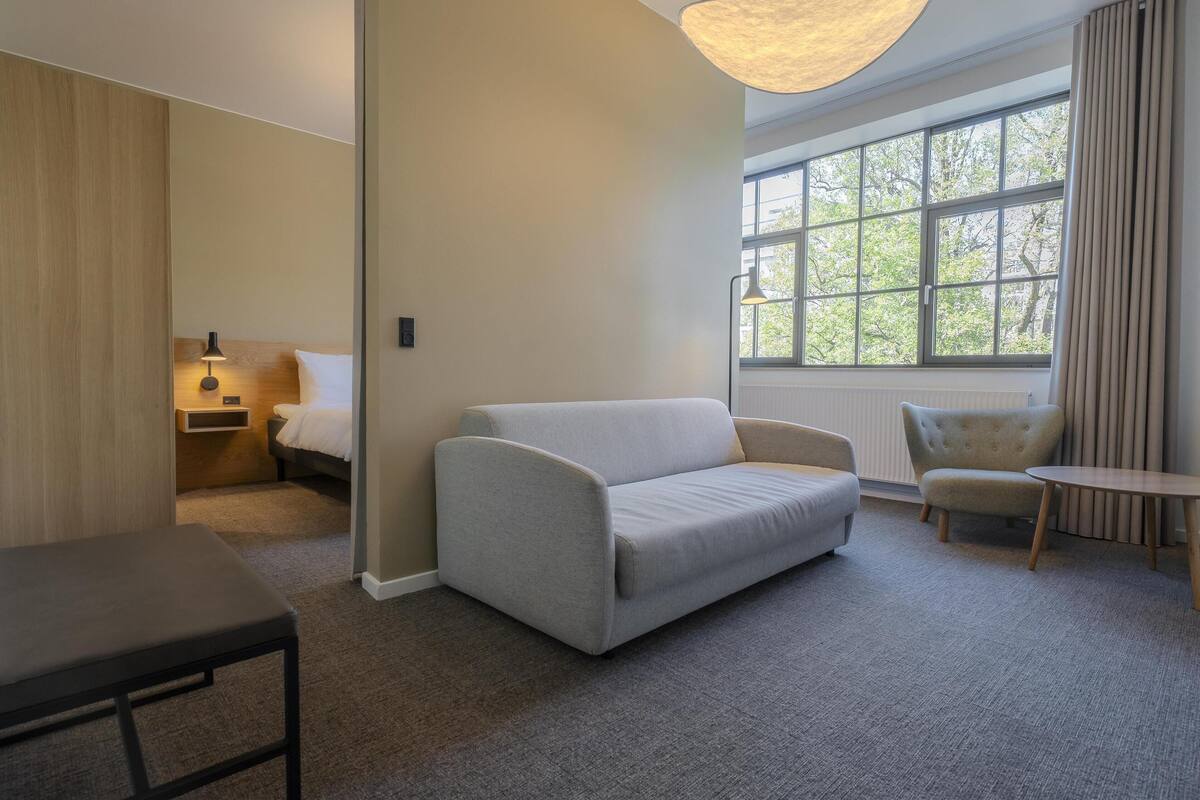 Et hotelværelse med en sofa, et lille bord, en stol, et natbord med lampe og et vindue med udsigt til træer.