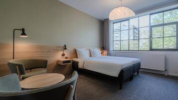 A modern hotel room with a bed, a desk, a chair, and a large window offering a view of buildings and trees.