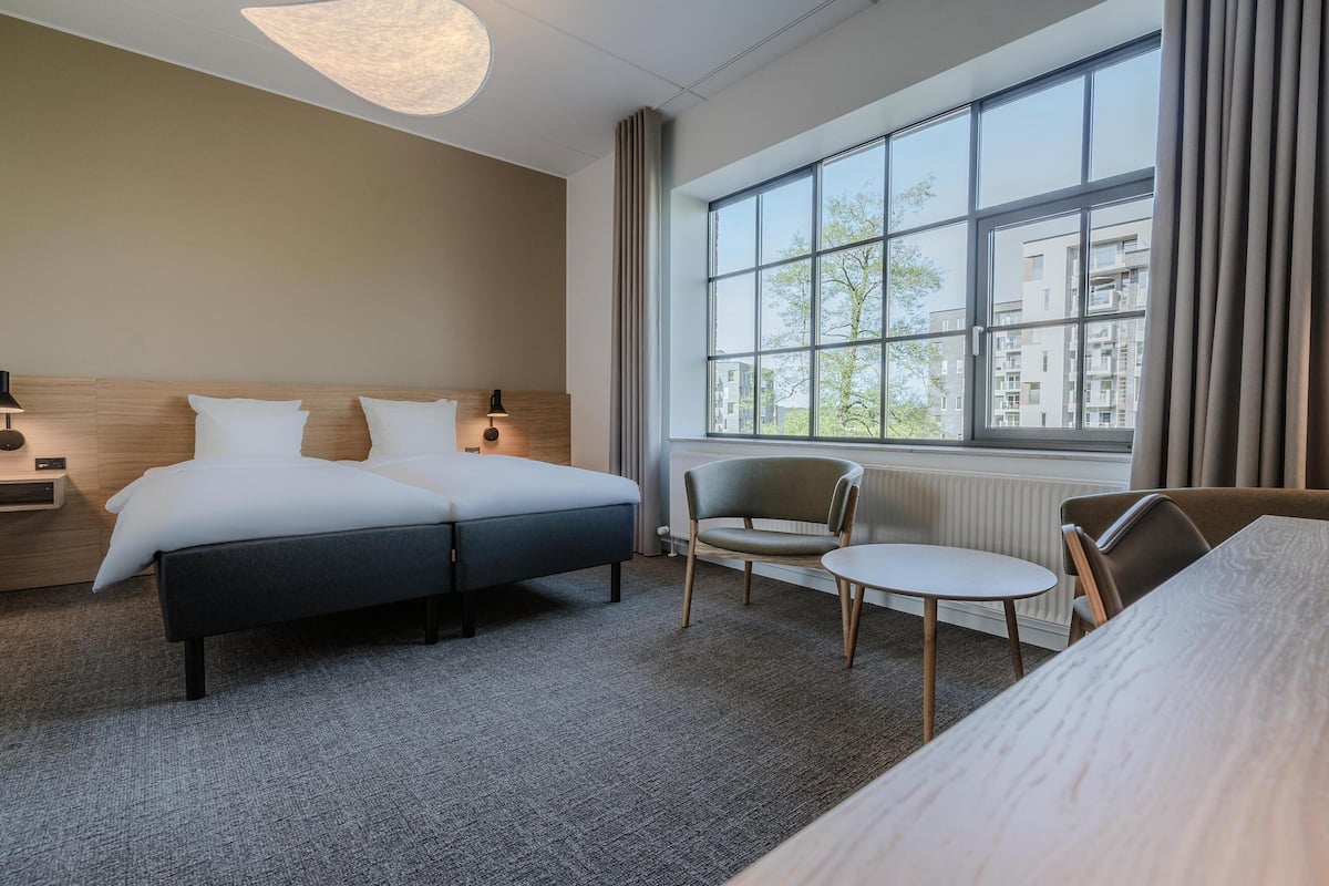 Et hotelværelse med dobbeltseng, et lille bord, to stole, en lampe og et stort vindue med udsigt til bygninger.