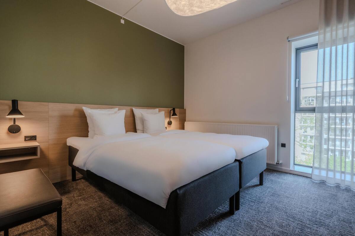 Et hotelværelse med en dobbeltseng, sengeborde, en bænk, en lampe og et stort vindue med gardiner.