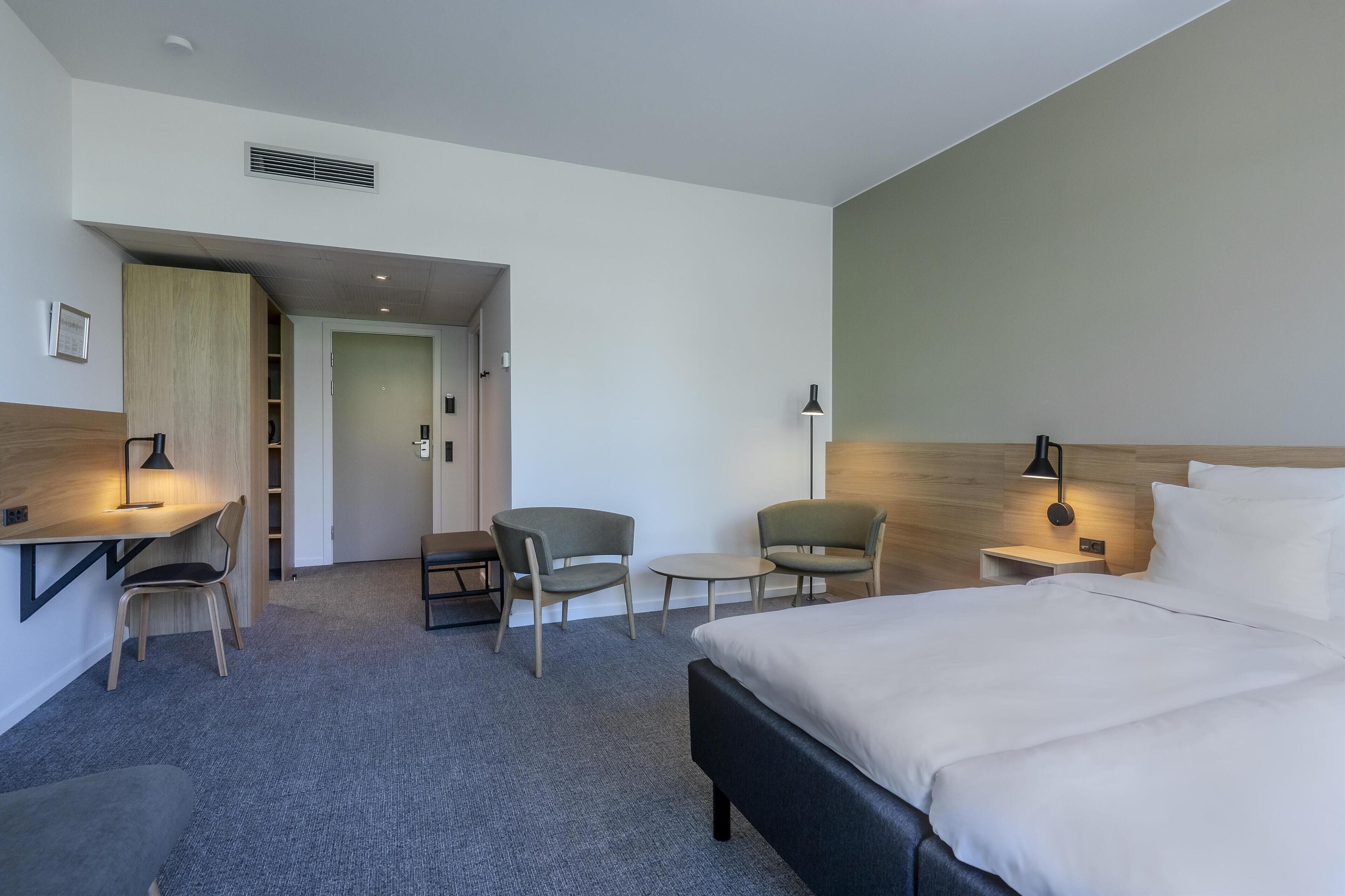 Habitación de hotel con una cama, dos sillas, un escritorio y una mesita pequeña. Se observa una puerta que da a otra habitación y un estante en la pared.