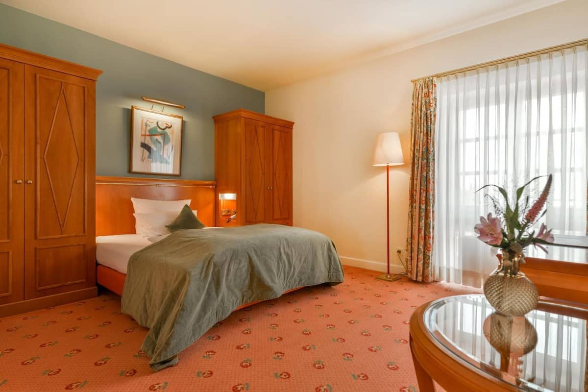 A hotel room with a bed, a wooden wardrobe, a painting on the wall, a floor lamp, and a window with curtains.