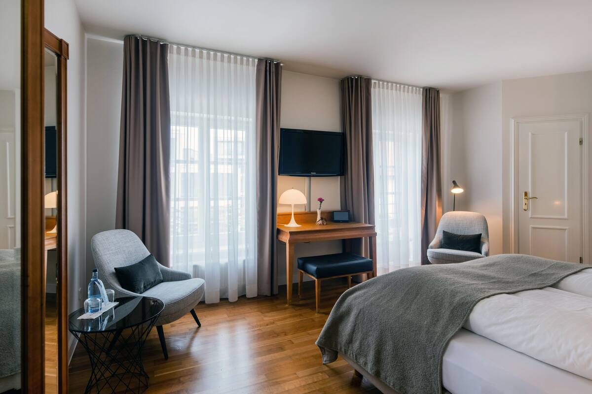 A hotel room with a bed, two armchairs, a desk with a TV, and a lamp.