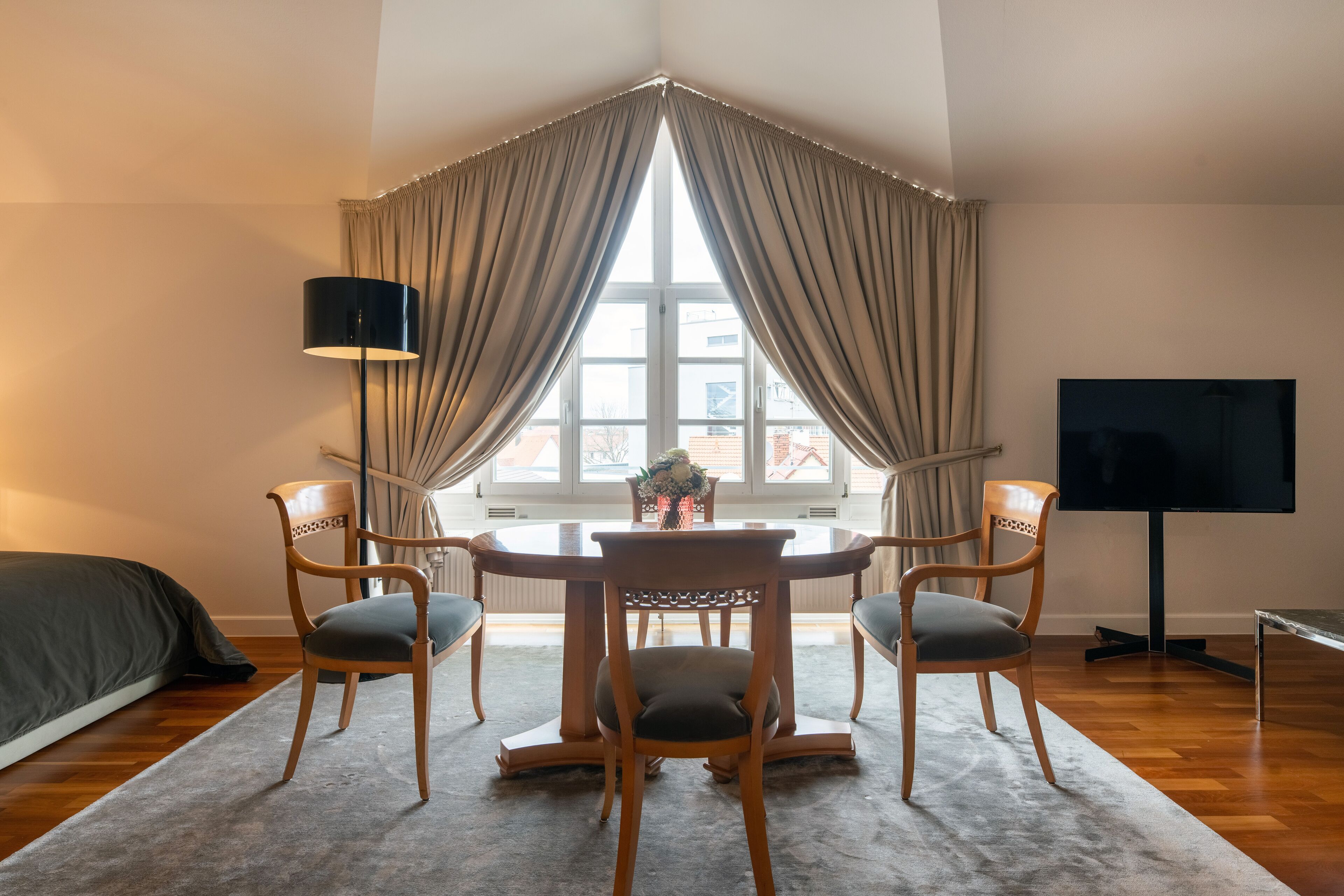 A corner room with a dining table, four chairs, a TV, and a window with curtains.
