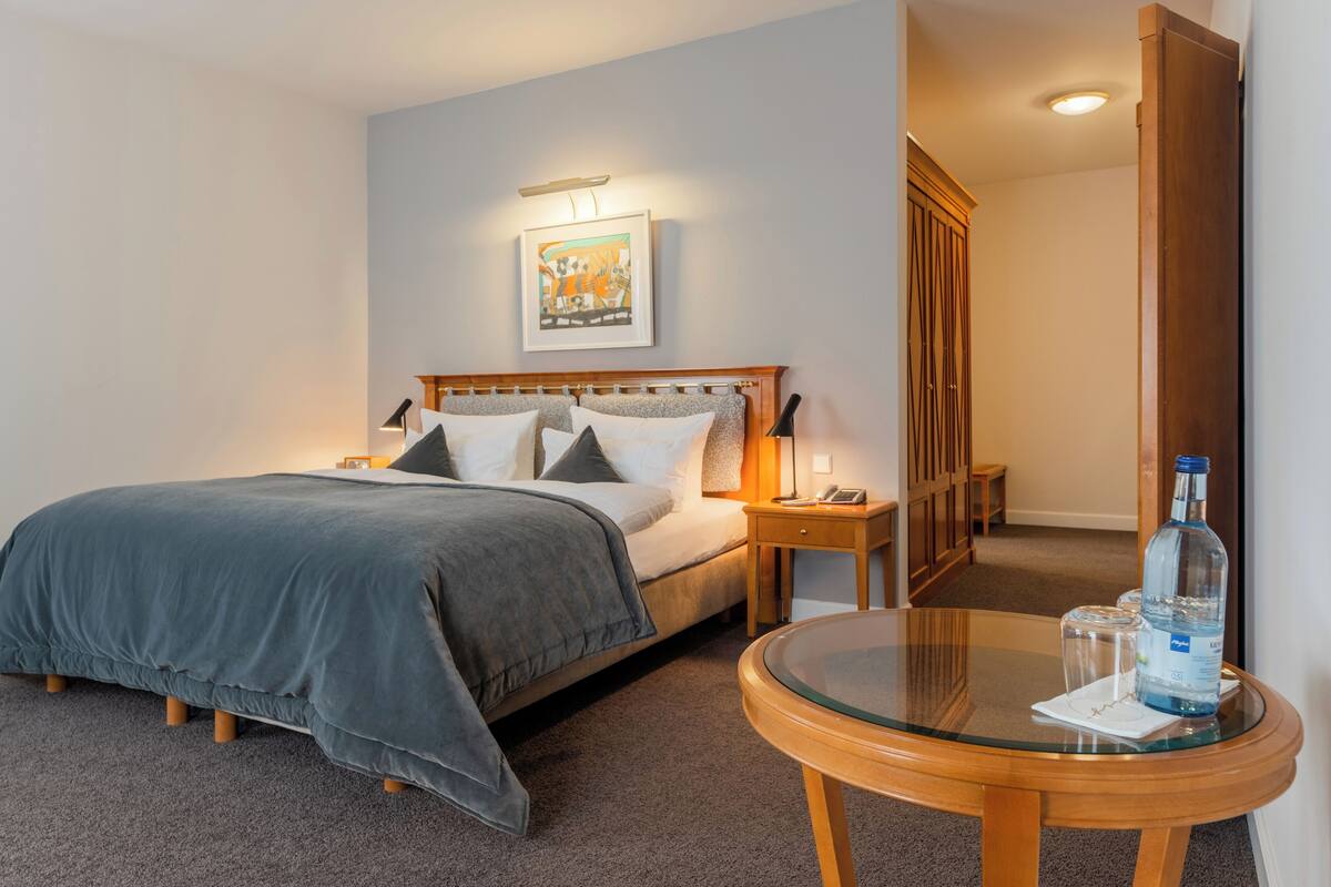 A neatly arranged hotel room with a bed, bedside table, and a round glass table with a bottle and glasses.
