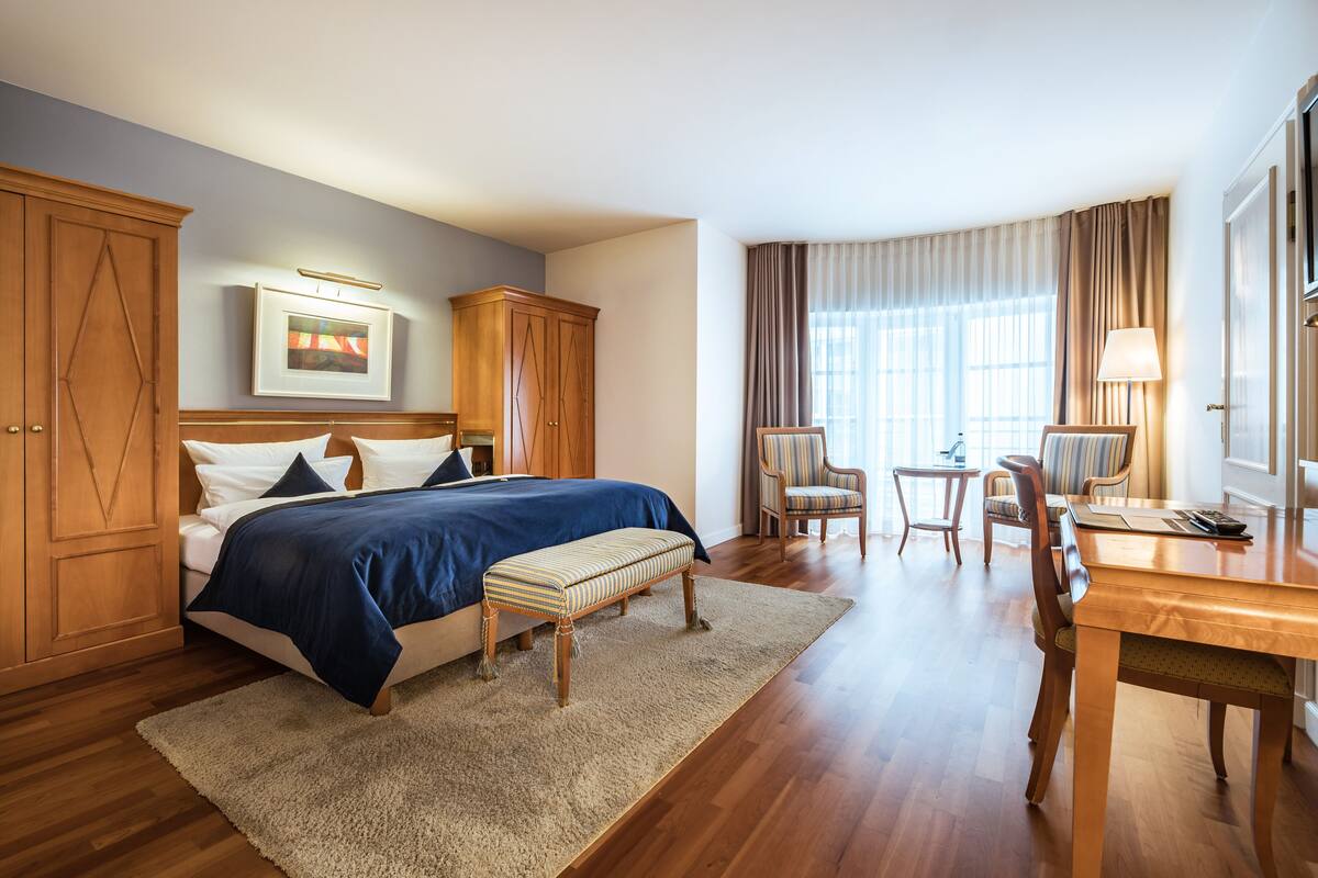 A hotel room with a large bed, wooden wardrobe, a desk with chairs, and a window with curtains.
