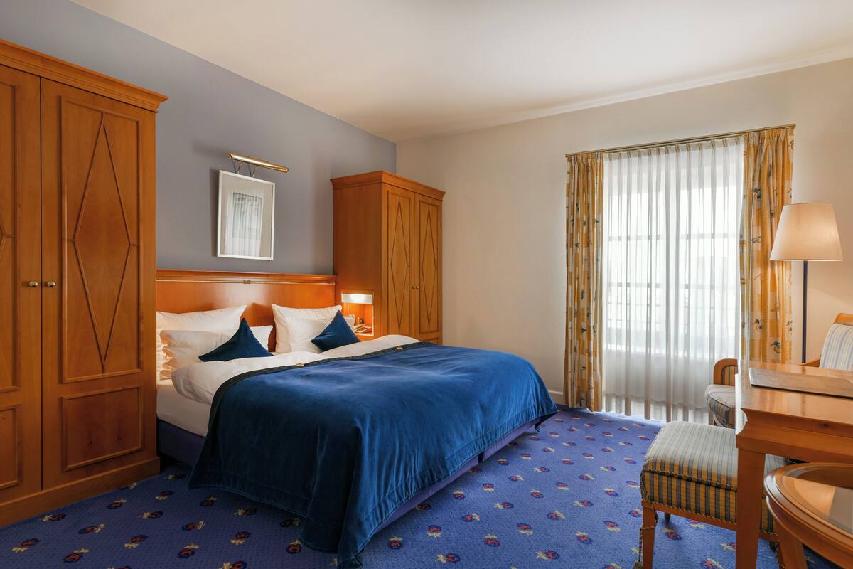 A hotel room with a bed, wooden wardrobe, a desk with a chair, and a window with curtains.