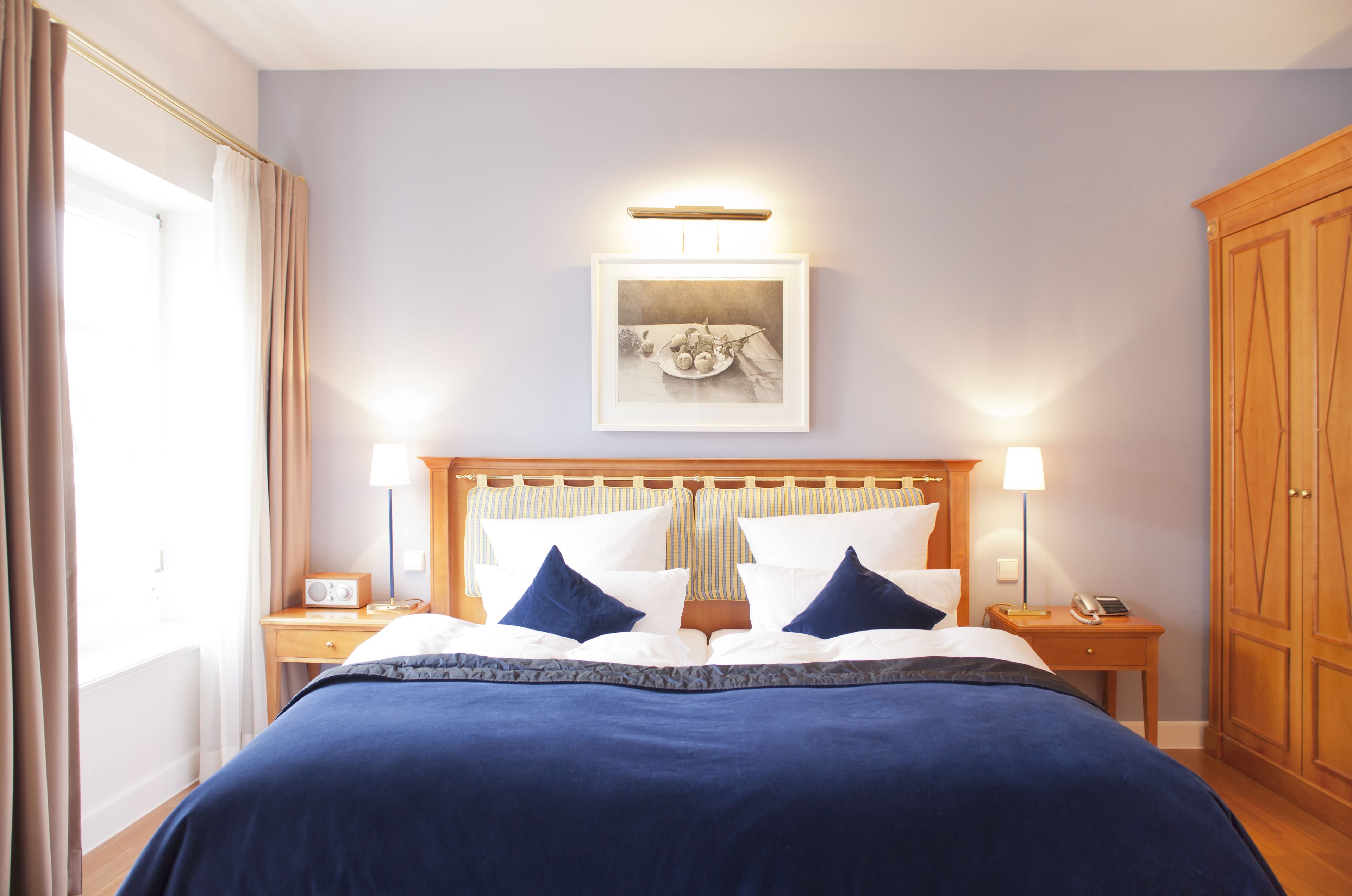 A hotel room with a large bed, two bedside tables, a nightstand, a wooden wardrobe, and a framed picture on the wall.