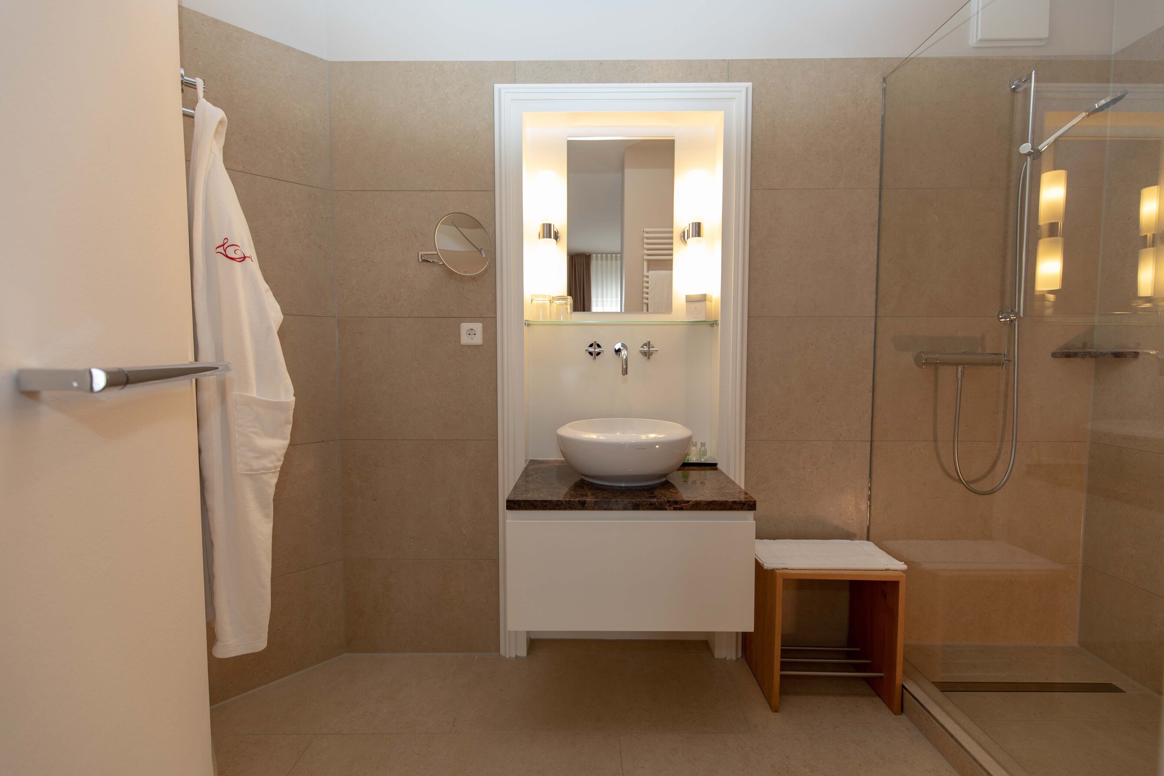 Un baño moderno con un lavabo blanco, una cabina de ducha con mampara de vidrio y un pequeño taburete de madera.