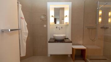Un baño moderno con un lavabo blanco, una cabina de ducha con mampara de vidrio y un pequeño taburete de madera.
