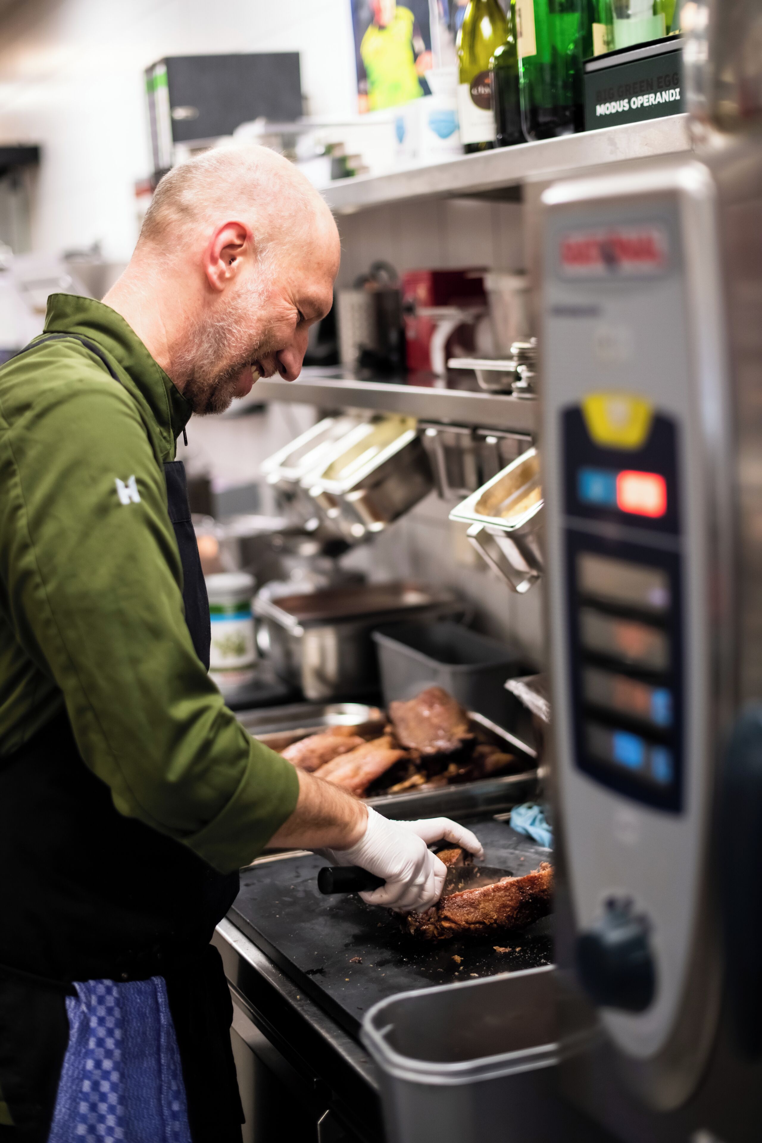 Eine Person in einer grünen Uniform bereitet Essen in einer gewerblichen Küche mit Edelstahlgeräten und einem Bedienfeld vor.