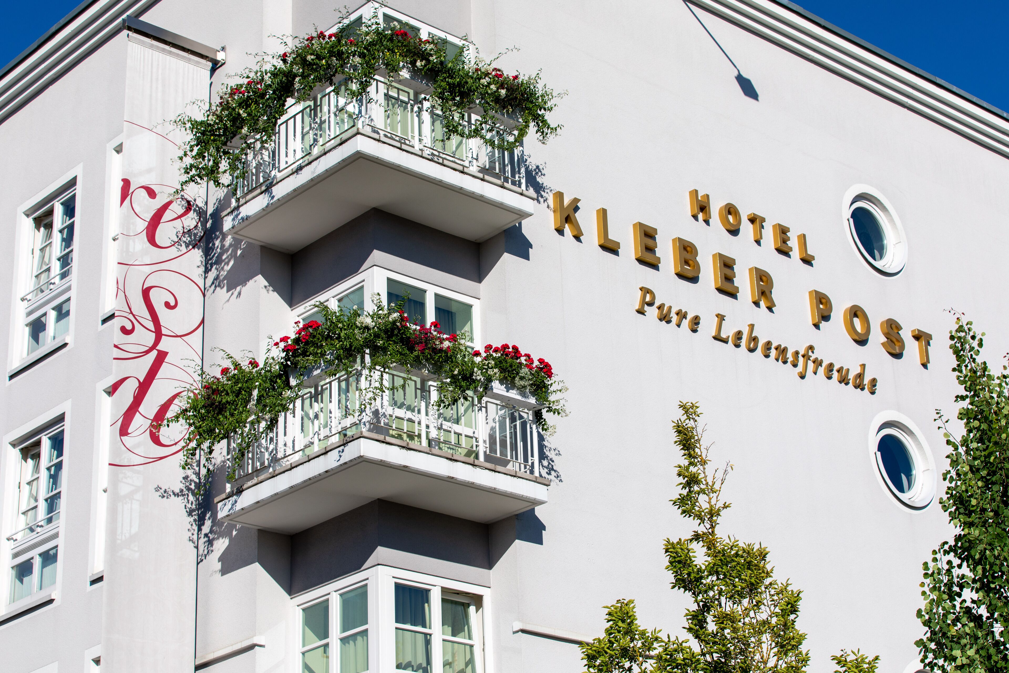 Ein Hotel namens 'Hotel Kleber Post' mit heller Fassade, Balkonen mit Blumenkästen und einem Schild mit dem Schriftzug 'Pure Lebensfreude'.