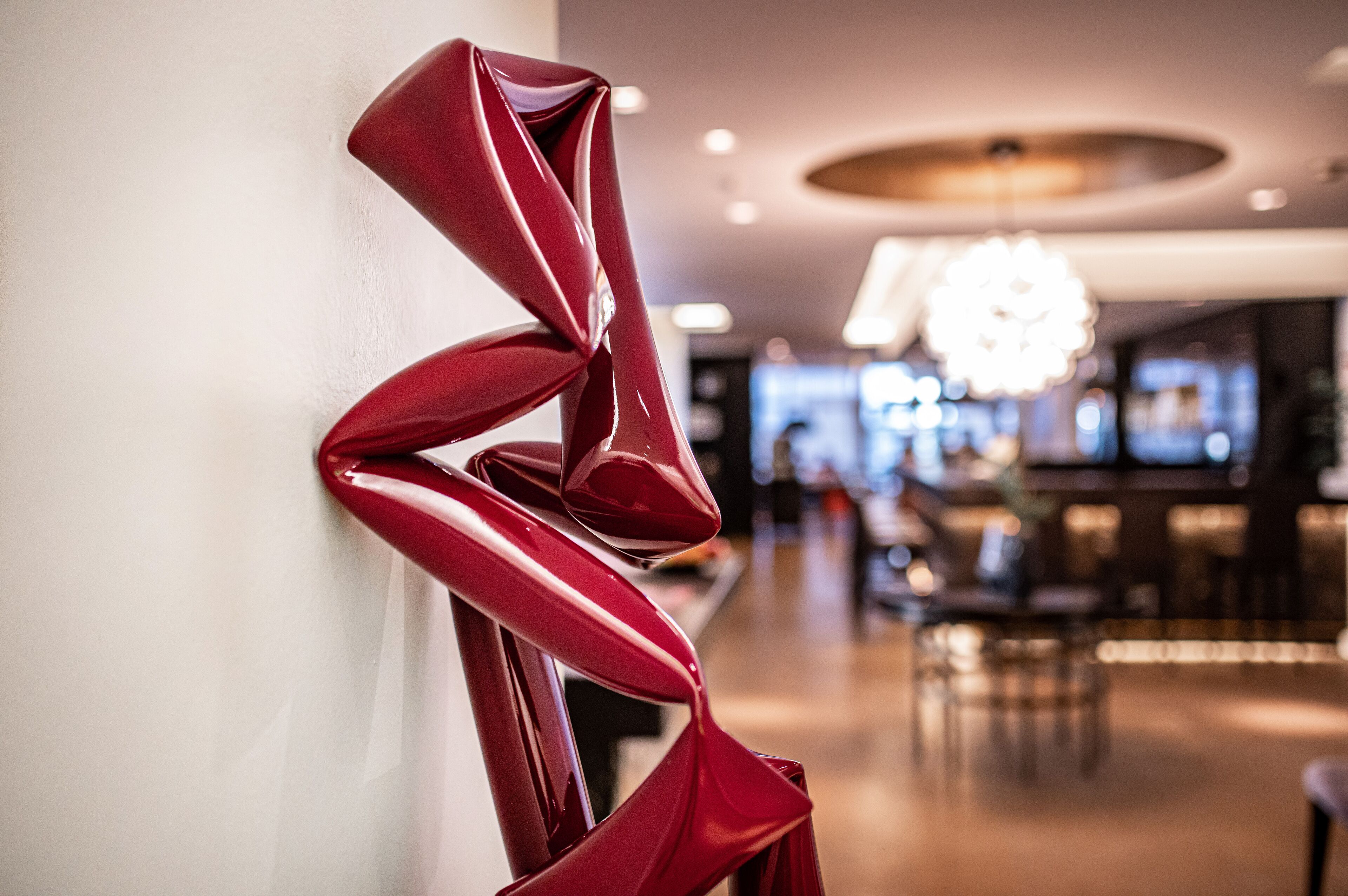 A modern interior with a red abstract sculpture on a wall, a bar area in the background, and a chandelier.