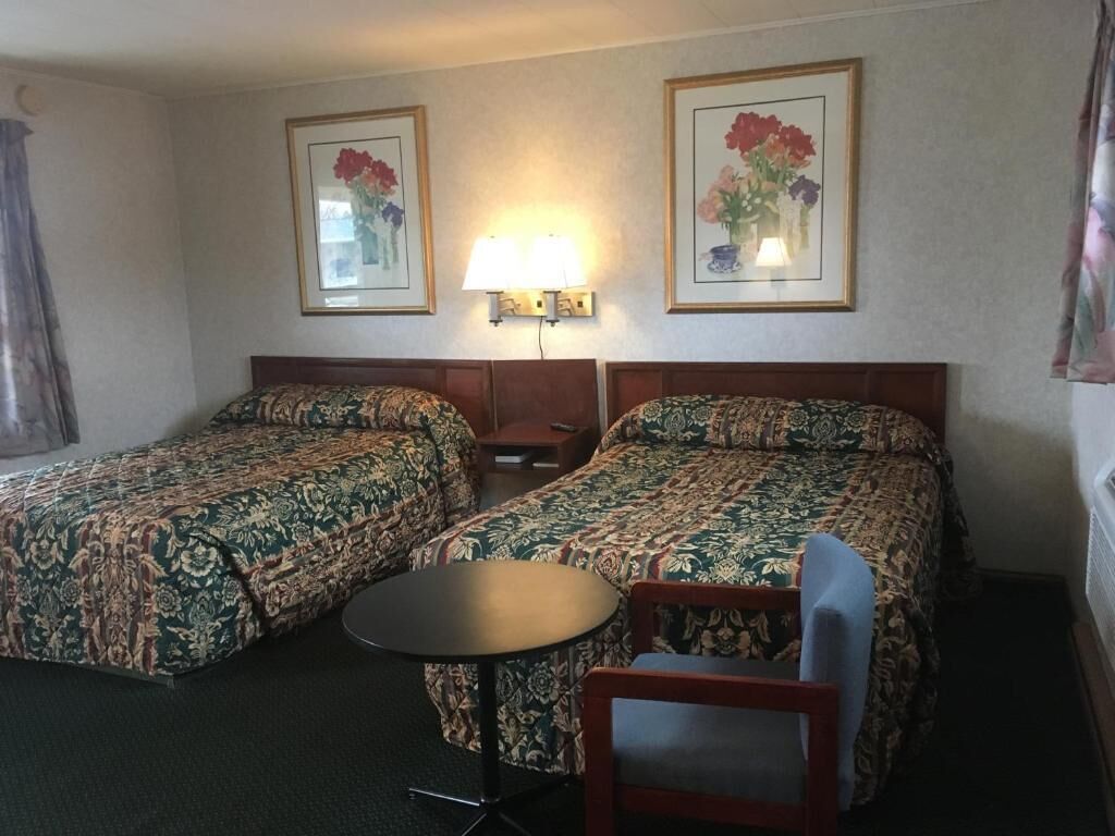 A hotel room with two beds, a small table, a chair, and a lamp.