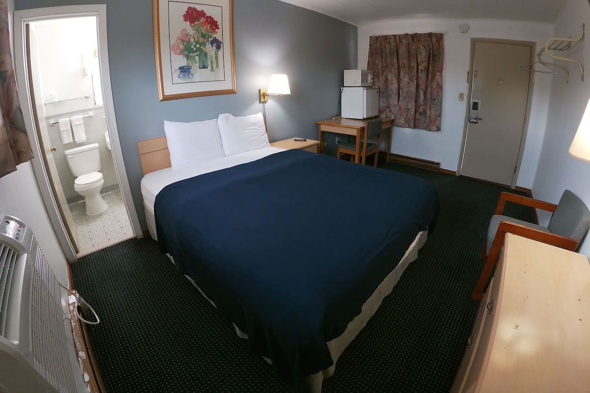 A hotel room with a bed, a desk, a chair, a lamp, a painting, a toilet, and a shower.