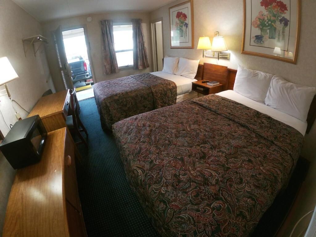 Quarto de hotel com duas camas, uma mesa, uma cadeira, uma televisão e uma janela com cortinas.