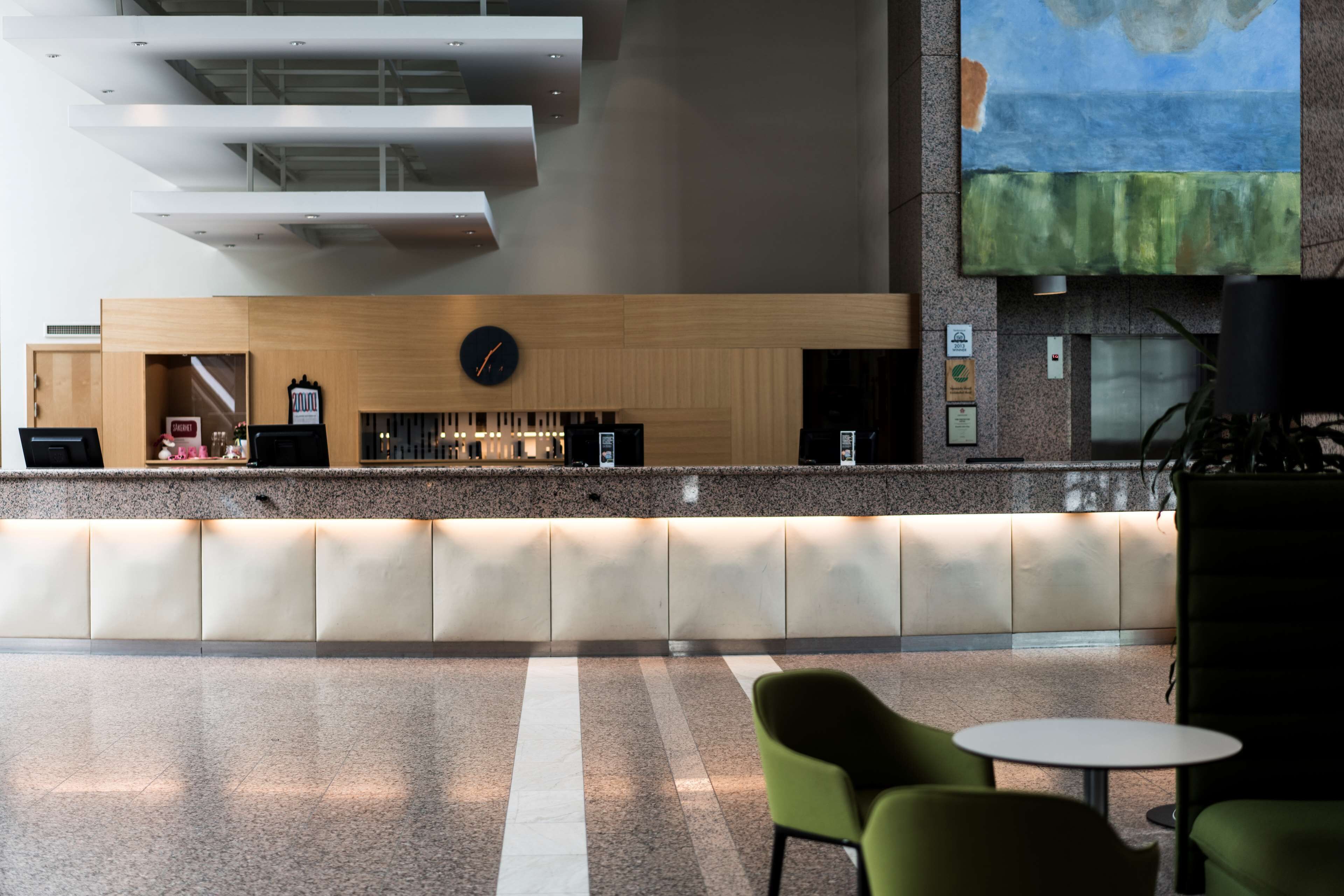 Ett modernt receptionsområde med en bardisk i trä, en klocka och en bar. I förgrunden finns gröna stolar och ett litet bord.