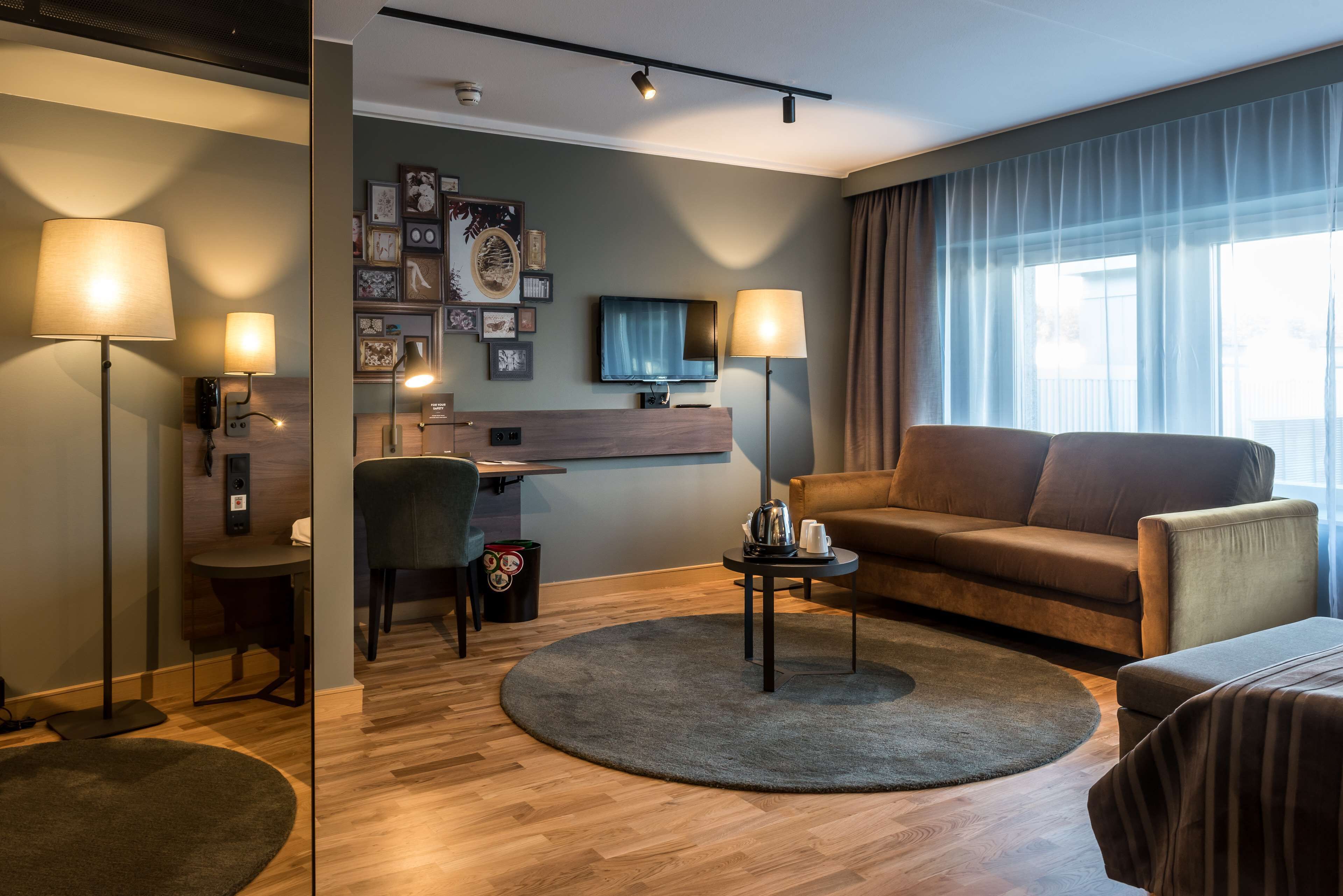Una habitación de hotel moderna con suelo de madera, un tapete redondo, un sofá, un escritorio con una silla, un televisor de pantalla plana y dos lámparas de pie.
