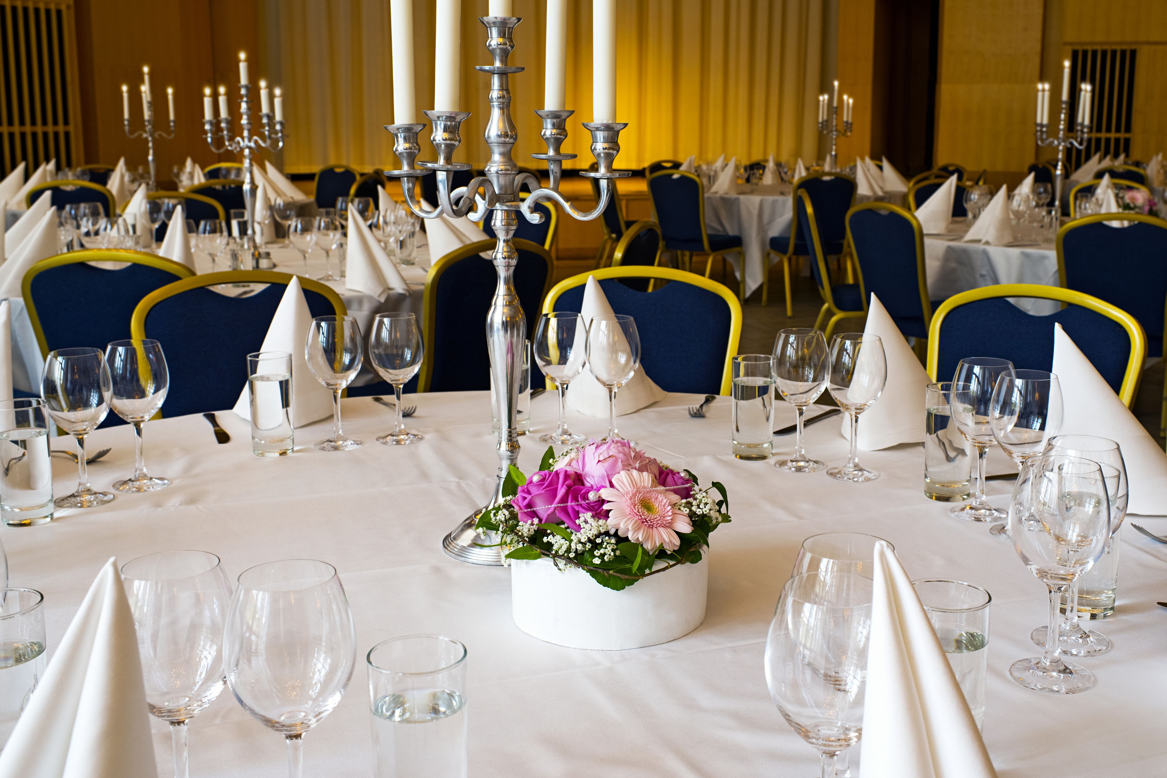 Un salón de banquetes con mesas redondas dispuestas para un evento formal, con manteles blancos, sillas azules y una decoración floral central.