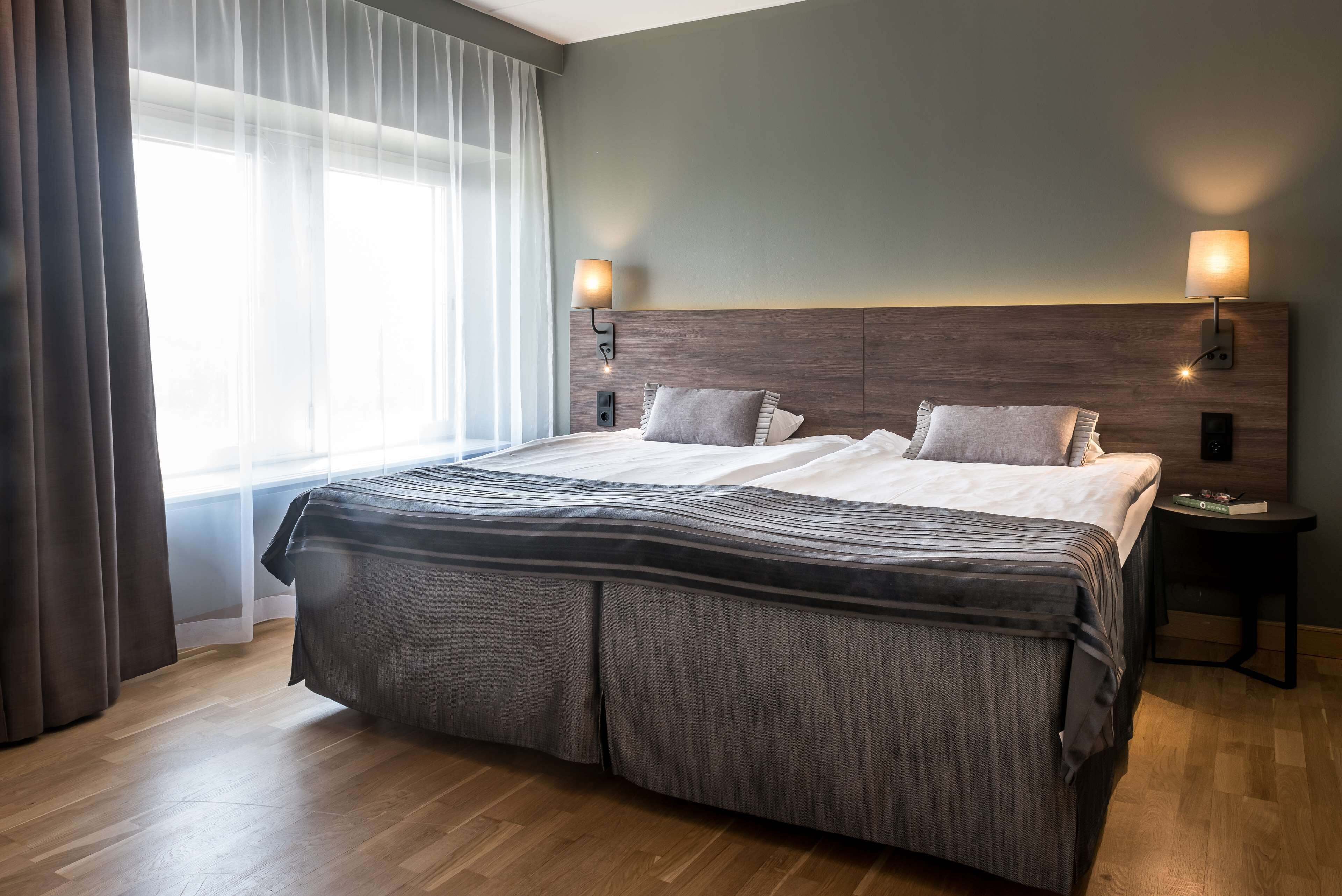 A hotel room with a large bed, two bedside lamps, a nightstand, and a window with sheer curtains.