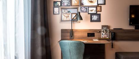 A hotel room with a desk, a chair, a bed, a TV, and a wall decorated with framed pictures.