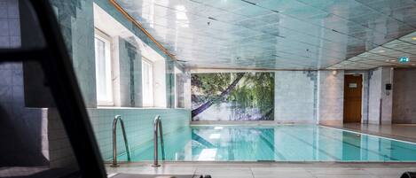 An indoor swimming pool with clear glass walls, a view of trees outside, and a tiled floor.
