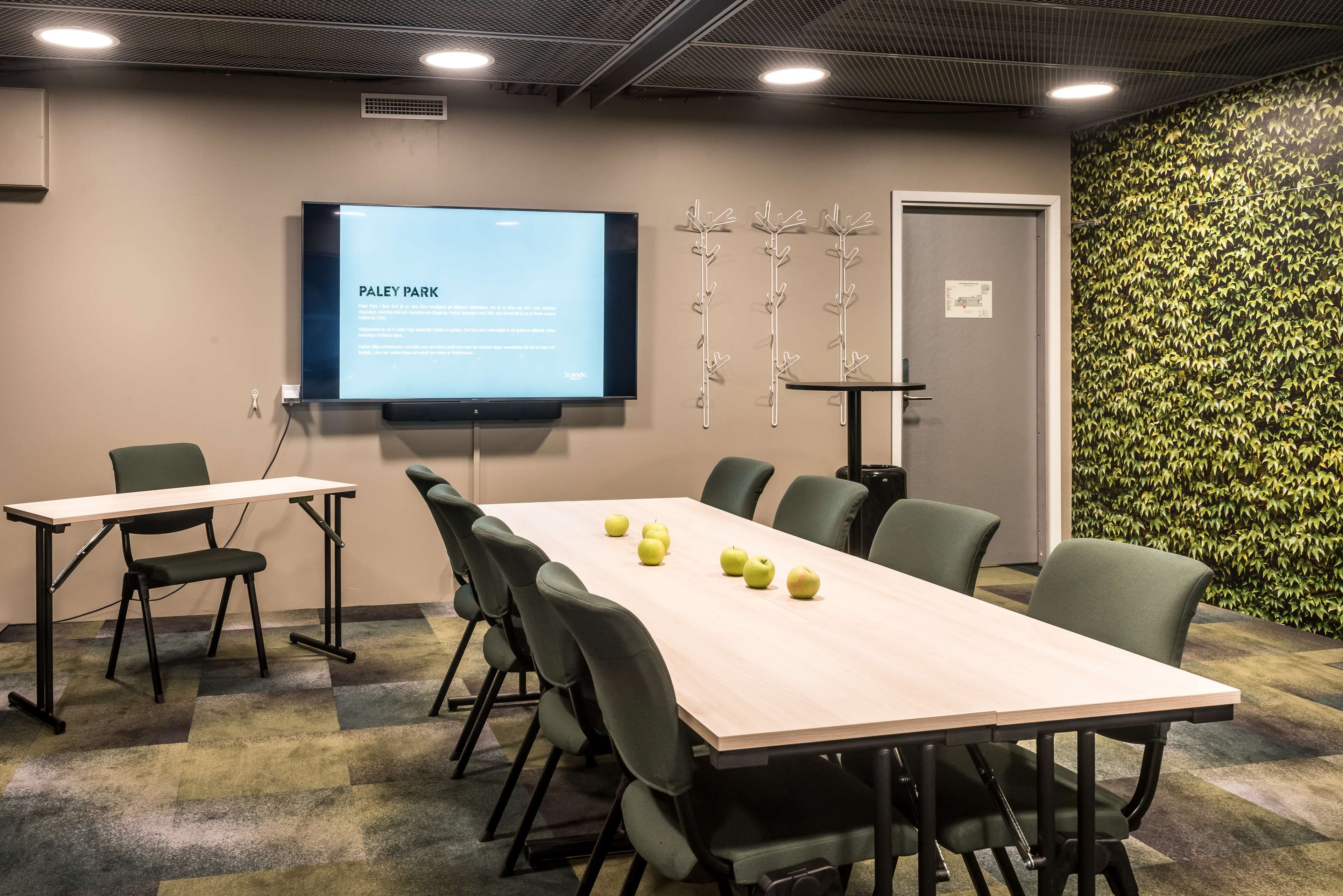Un salón de conferencias con una gran mesa, sillas, una pantalla de proyector y una decoración en la pared.