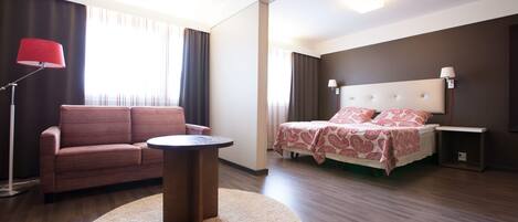 A modern hotel room with a sofa, a round table, a bed with patterned bedding, and a nightstand.