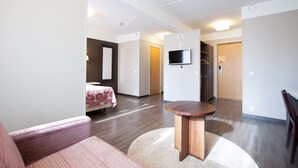 Une chambre d’hôtel moderne, équipée d’un lit, d’une table ronde en bois, d’un téléviseur fixé au mur et d’un couloir menant à d’autres chambres.