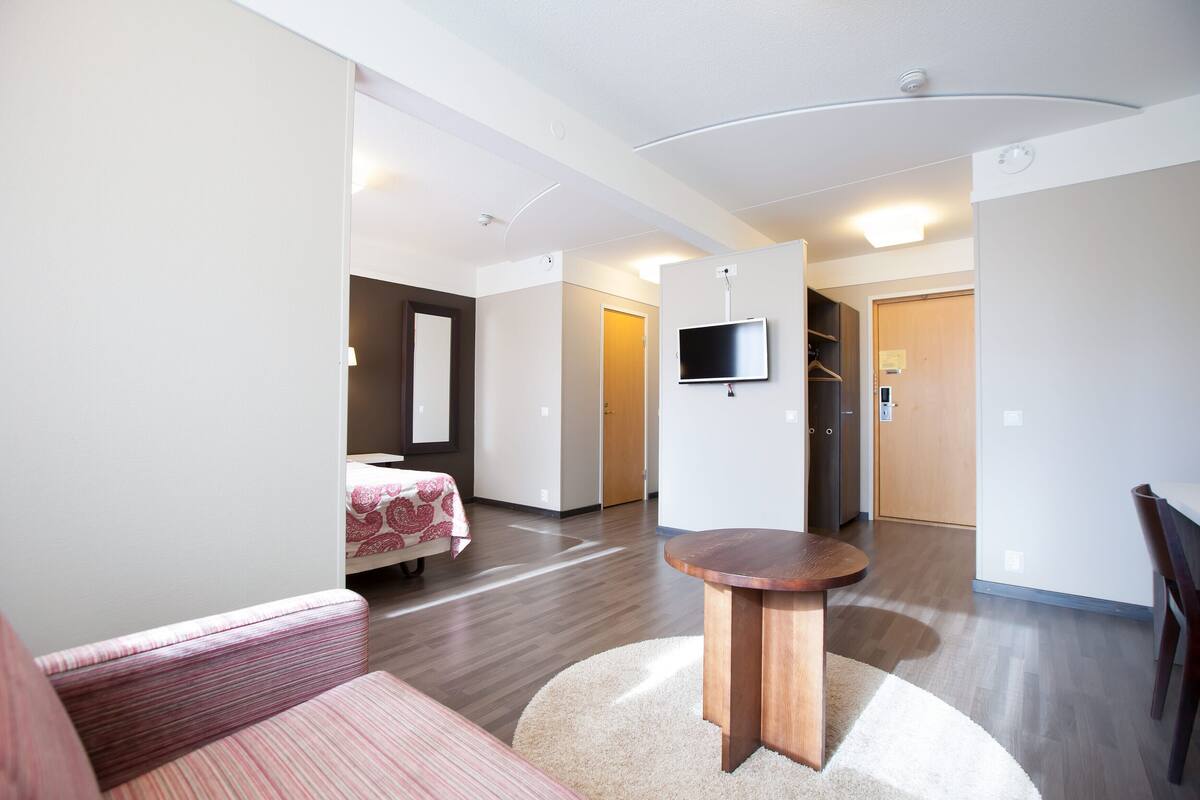 A modern hotel room with a bed, a round wooden table, a TV mounted on the wall, and a hallway leading to other rooms.
