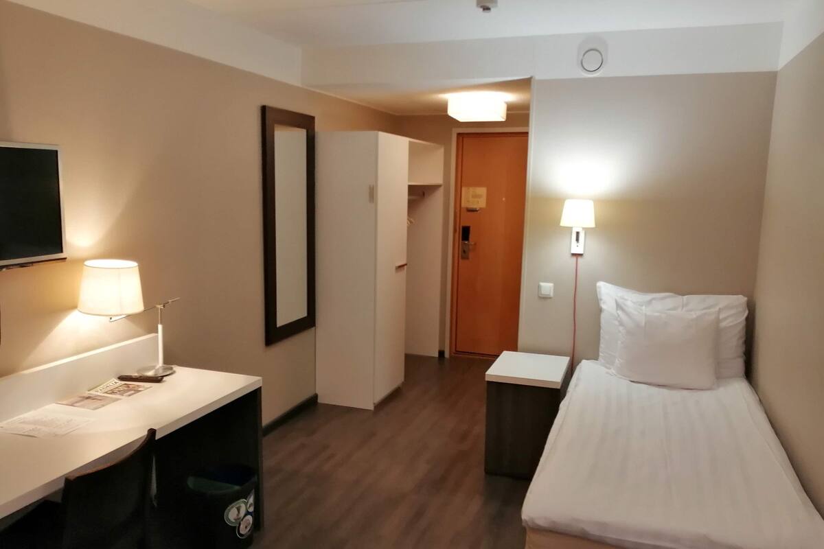 A hotel room with a bed, a desk, a chair, a trash bin, a TV, and a door.