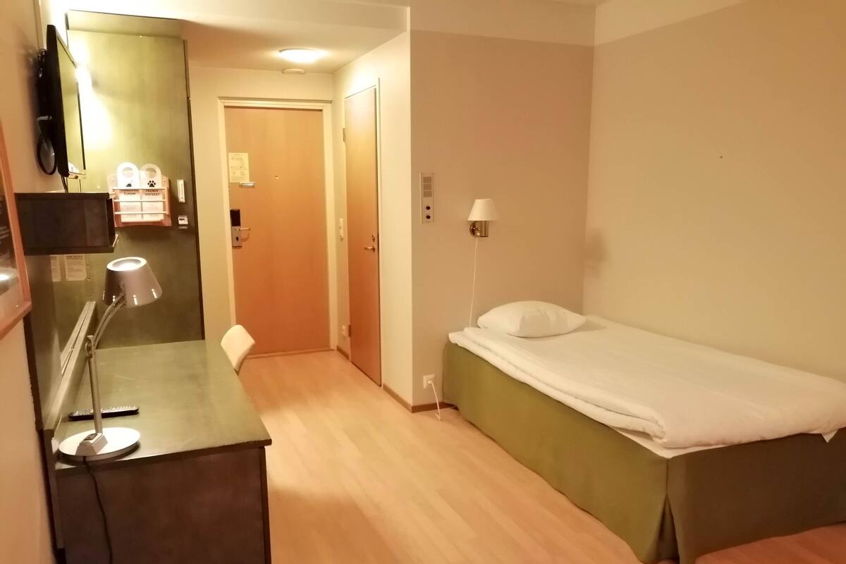 A hotel room with a bed, a desk with a lamp, a chair, and a small kitchenette area.