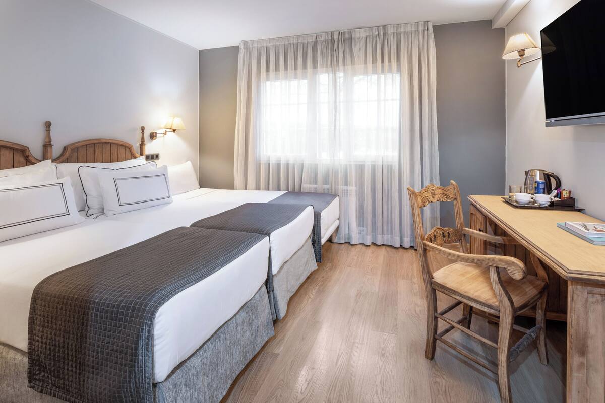A hotel room with a large bed, two bedside tables, a desk with a chair, a television, and a window with sheer curtains.